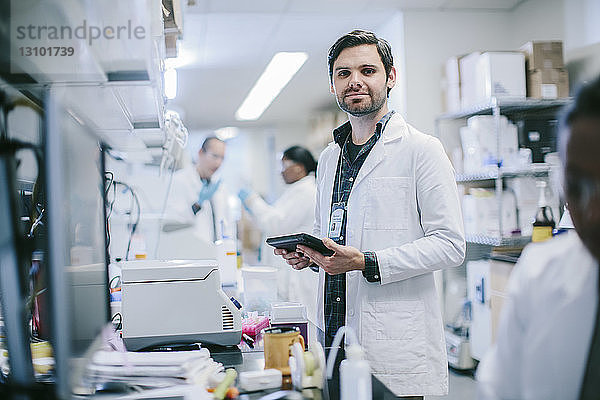 Porträt eines männlichen Arztes mit Tablet-Computer und Mitarbeitern  die im Krankenzimmer arbeiten