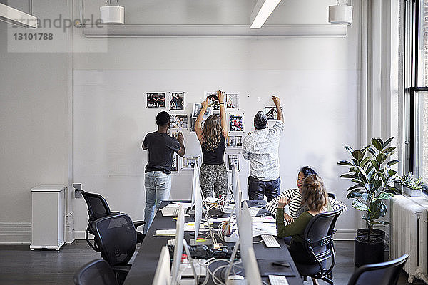 Geschäftsleute befestigen Fotoausdrucke am Schwarzen Brett  während weibliche Kollegen im Büro am Konferenztisch sitzen