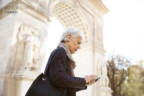 Seitenansicht einer älteren Frau  die ein Smartphone benutzt  während sie in der Stadt gegen den Triumphbogen steht