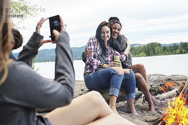 Frau fotografiert glückliche Freundinnen  die am Lagerfeuer am Fluss auf einem Baumstamm sitzen