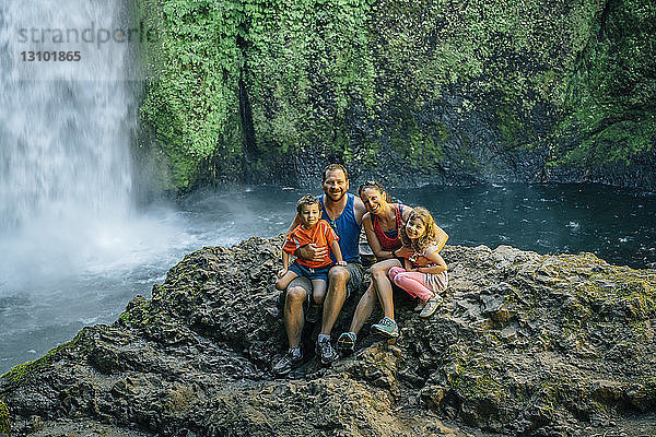 Porträt einer glücklichen Familie  die auf Felsen vor einem Wasserfall im Wald sitzt