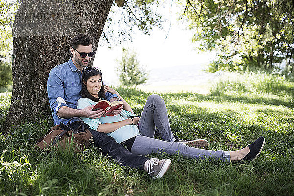 Paar liest Buch beim Entspannen am Baum im Park