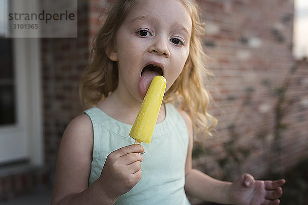 Porträt eines Mädchens  das Eis am Stiel isst  während es gegen ein Haus steht