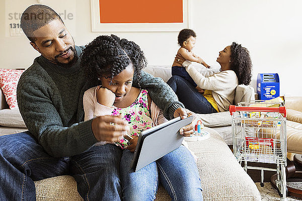 Vater und Tochter benutzen einen Tablet-Computer  während die Mutter mit dem Sohn auf dem Sofa spielt
