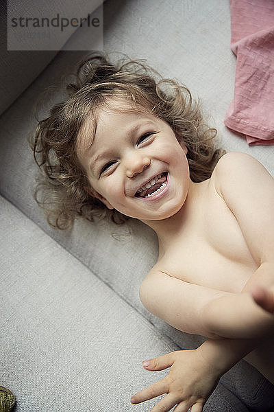 Hochwinkelporträt eines glücklichen Jungen ohne Hemd  der zu Hause auf dem Sofa liegt