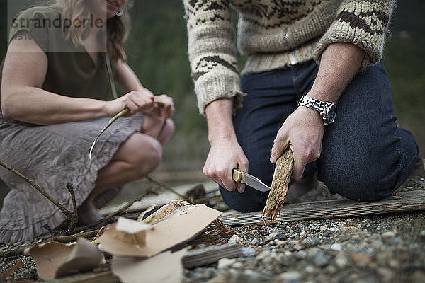 Freund schneidet in der Mitte Holz  während seine Freundin auf dem Campingplatz kniet