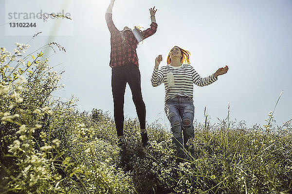 Verspielte Freundinnen springen im Sommer auf Grasfeld gegen klaren Himmel