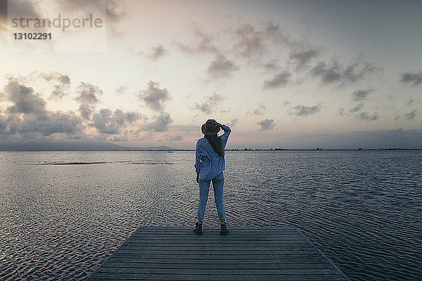 Rückansicht einer Frau  die bei Sonnenuntergang auf dem Pier über dem Meer gegen den Himmel steht