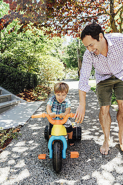 Glücklicher Vater hilft Sohn beim Dreiradfahren im Park