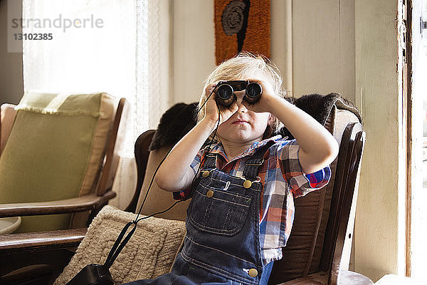 Junge schaut durch ein Fernglas  während er zu Hause auf einem Stuhl sitzt