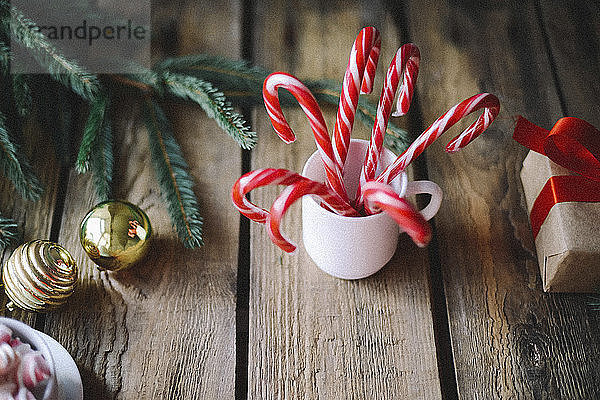 Nahaufnahme von Zuckerstangen in Tasse durch Kugeln und Zweig auf Holztisch zu Weihnachten