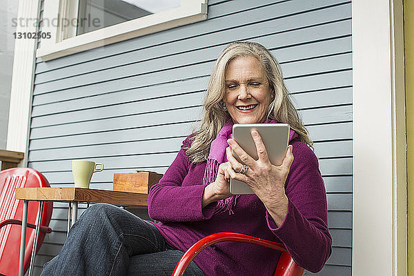 Lächelnde ältere Frau benutzt Tablet-Computer  während sie auf einem Stuhl auf der Veranda sitzt