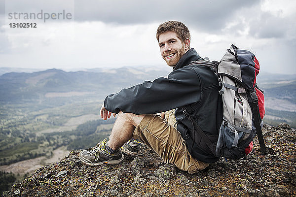 Porträt eines lächelnden Mannes  der einen Rucksack trägt  während er auf einem Berg sitzt