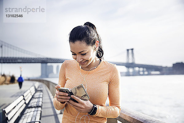 Glückliche Sportlerin hört Musik über Smartphone mit Williamsburg Bridge im Hintergrund