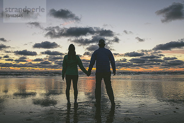 Rückansicht eines Paares  das am Long Beach vor bewölktem Himmel Händchen hält