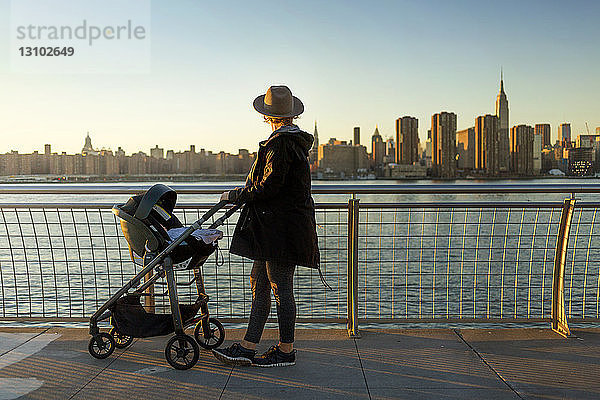 Seitenansicht einer Frau  die die Skyline der Stadt betrachtet  während sie bei Sonnenuntergang mit einem Kinderwagen auf der Promenade steht