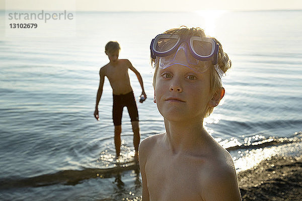 Porträt eines Jungen mit Schwimmbrille am Ufer mit einem Freund im Hintergrund
