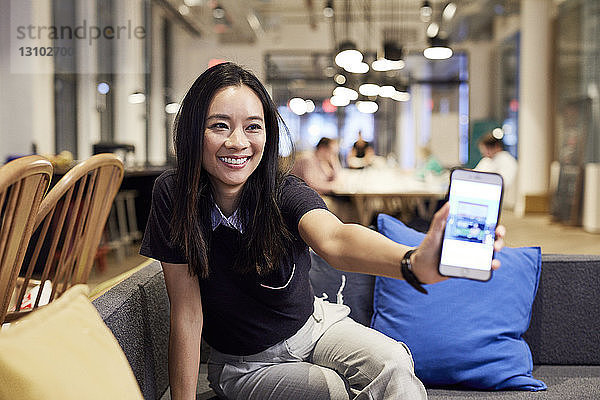 Lächelnde Geschäftsfrau zeigt Mobiltelefon  während sie im Büro auf dem Sofa sitzt