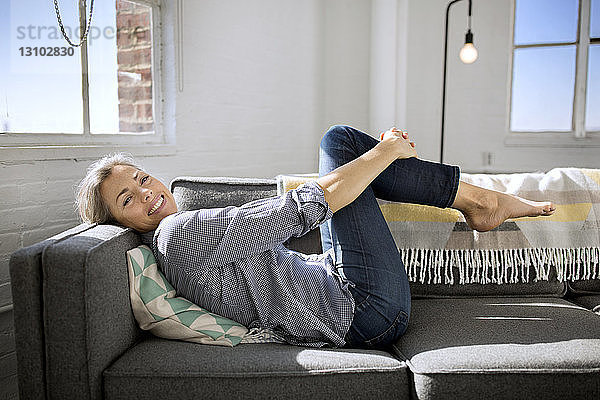 Porträt einer glücklichen Frau  die die Knie umarmt  während sie zu Hause auf dem Sofa liegt