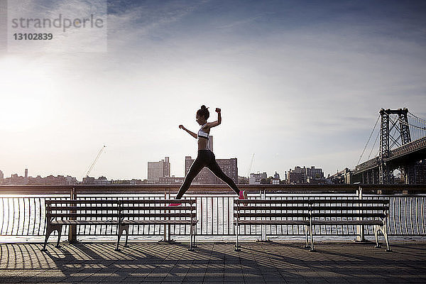 Sportliche Frau springt auf Bänken mit der Williamsburg Bridge im Hintergrund