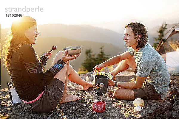 Glückliches Paar isst  während es auf einem Berg vor klarem Himmel sitzt