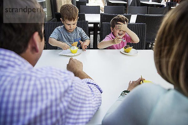 Glücklicher Junge bedeckt die Augen  während er mit seiner Familie im Restaurant Dessert isst