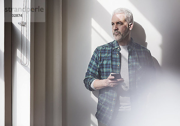 Nachdenklicher  reifer Mann hält Smartphone in der Hand und schaut zu Hause durchs Fenster