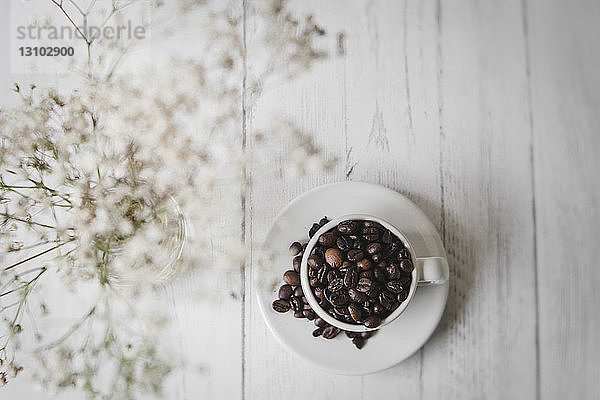 Draufsicht auf Kaffeebohnen in Tasse mit Untertasse durch Blumenvase auf Holztisch