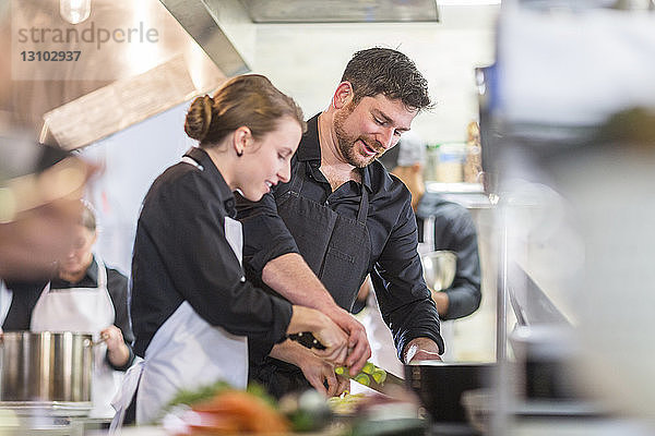 Männlicher Chefkoch unterstützt weibliche Mitarbeiterin beim Gemüseschneiden in der Restaurantküche