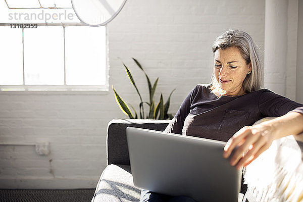 Frau benutzt Laptop  während sie zu Hause auf dem Sofa im Wohnzimmer sitzt