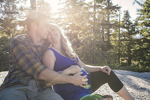 Mann berührt den Bauch einer schwangeren Frau  während er an einem sonnigen Tag an Bäumen sitzt
