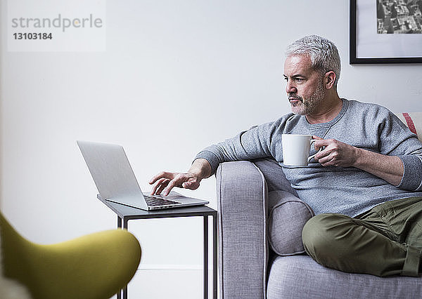 Reifer Mann benutzt Laptop  während er zu Hause Kaffeetasse hält