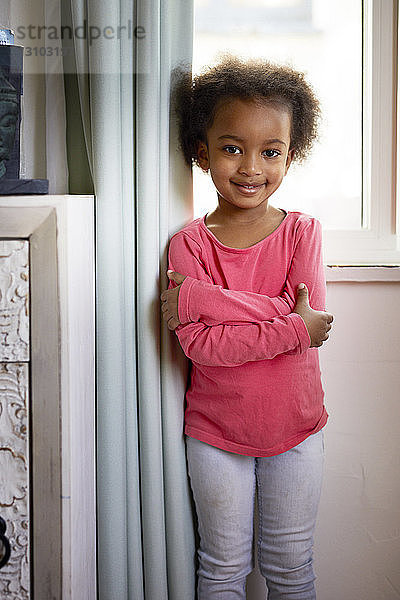 Porträt eines lächelnden Mädchens mit verschränkten Armen  das zu Hause vor einem Vorhang steht