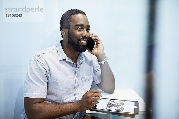 Lächelnder Geschäftsmann telefoniert mit einem Mobiltelefon  während er ein digitales Tablet in der Bürokabine benutzt