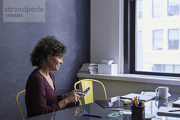 Lächelnde Geschäftsfrau benutzt Mobiltelefon  während sie im Büro am Konferenztisch sitzt
