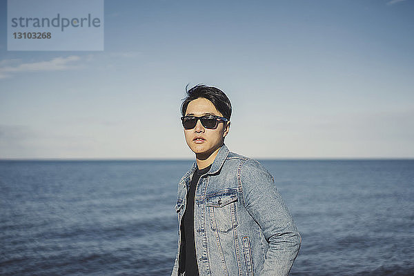 Junger Mann trägt Sonnenbrille und Jeansjacke  während er gegen Meer und Himmel steht