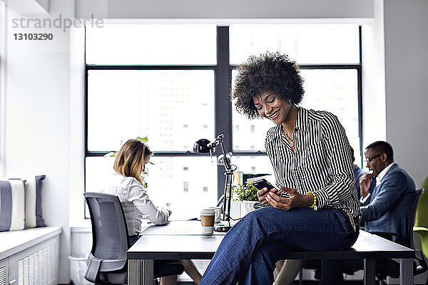 Glückliche Geschäftsfrau benutzt Smartphone  während Kollegen im Hintergrund arbeiten