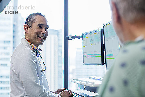 Lächelnder männlicher Arzt im Gespräch mit Patient  während er den Computer auf der Krankenhausstation benutzt