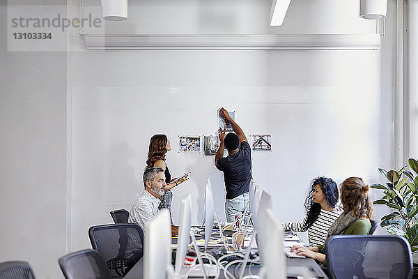 Geschäftsleute diskutieren am Konferenztisch  während Kollegen Fotoausdrucke am Schwarzen Brett im Büro anbringen