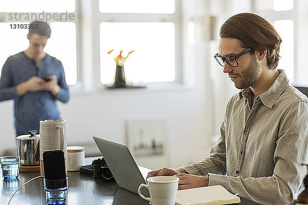 Mann benutzt zu Hause einen Laptop-Computer mit seinem Freund im Hintergrund