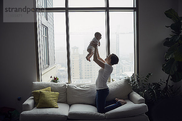 Seitenansicht einer glücklichen Mutter  die ihren Sohn hochhebt  während sie zu Hause auf dem Sofa am Fenster kniet