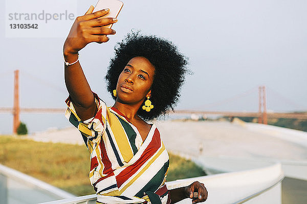 Selbstbewusste junge Frau macht ein Selfie mit der Brücke 25 de Abril im Hintergrund  Lissabon  Portugal