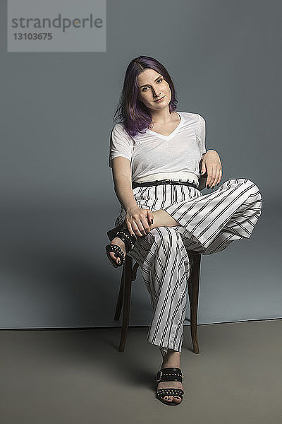 Porträt selbstbewusste und modische junge Frau sitzt auf einem Stuhl vor grauem Hintergrund