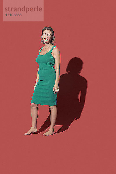 Porträt lächelnde  selbstbewusste barfüßige Frau in grünem Kleid vor rotem Hintergrund