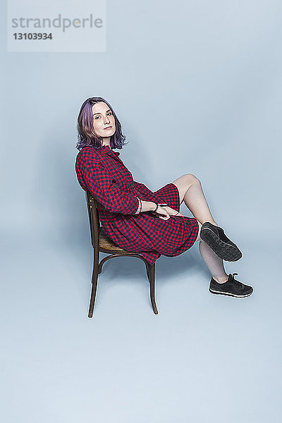 Porträt selbstbewusste und modische junge Frau sitzt auf einem Stuhl vor blauem Hintergrund