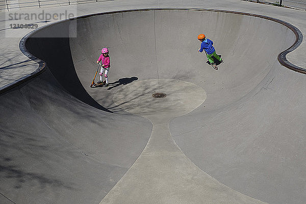 Bruder und Schwester auf Motorrollern spielen im sonnigen Skatepark