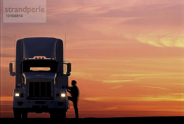 Die Silhouette eines Lkw-Fahrers  der bei Sonnenaufgang in das Fahrerhaus seiner gewerblichen Zugmaschine der Klasse 8 einsteigt.