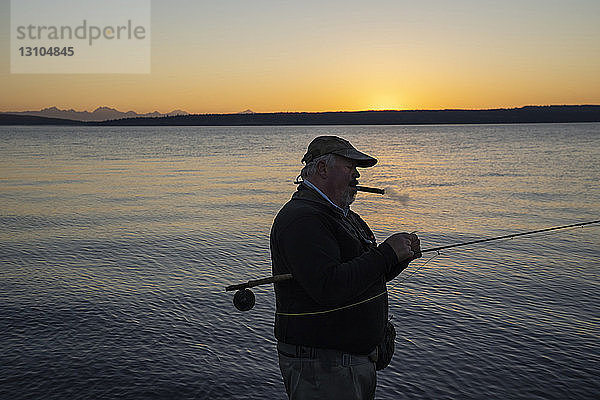 Ein älterer kaukasischer Fliegenfischer genießt eine Zigarre bei Sonnenaufgang  bevor er an der Westküste der USA mit der Fliege auf Cutthroat-Forelle und Lachs fischt