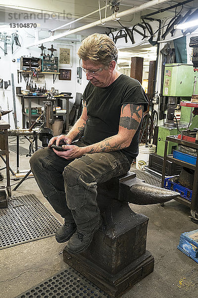 Männlicher Schmied  der sein Telefon auf Nachrichten überprüft  während er auf einem Amboss in seinem Atelier sitzt.