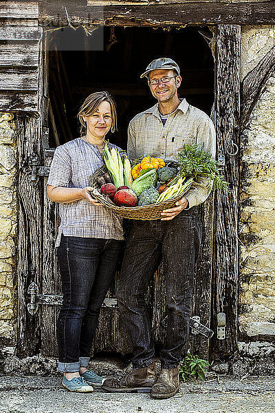 Lächelnder Bauer und Frau stehen im Scheunentor  halten einen Korb mit frisch geerntetem Gemüse und schauen in die Kamera.